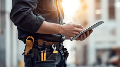 A focused and professional facilities management technician, wearing a uniform and toolbelt, holding a tablet where he's utilizing field service software to manage the work order.