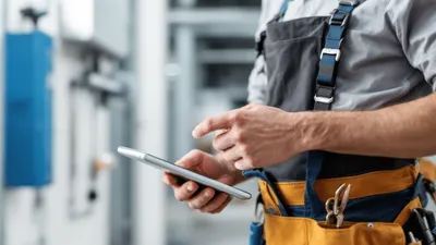 A focused and professional construction field service technician, wearing a uniform and toolbelt, holding a tablet where he's utilizing field service software to manage the work order.